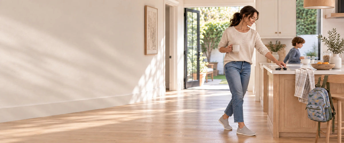 Woman with a mug of coffee crossing a sunny kitchen at home while her son eats breakfast, wearing Ash Gray COOPS Low Top sock shoes