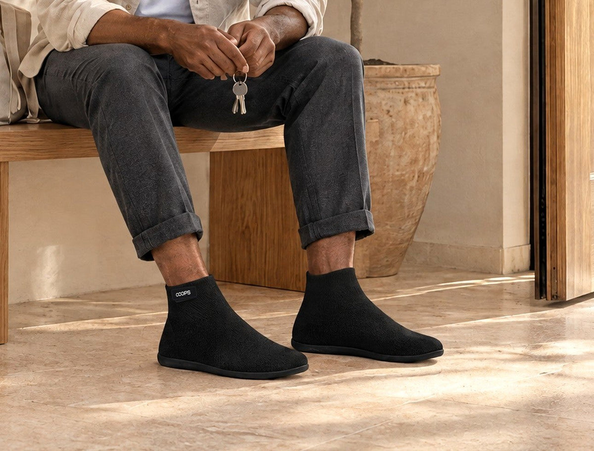 A man on an oak entry bench holding his keys by the open front door, wearing Black COOPS High Top sock shoes