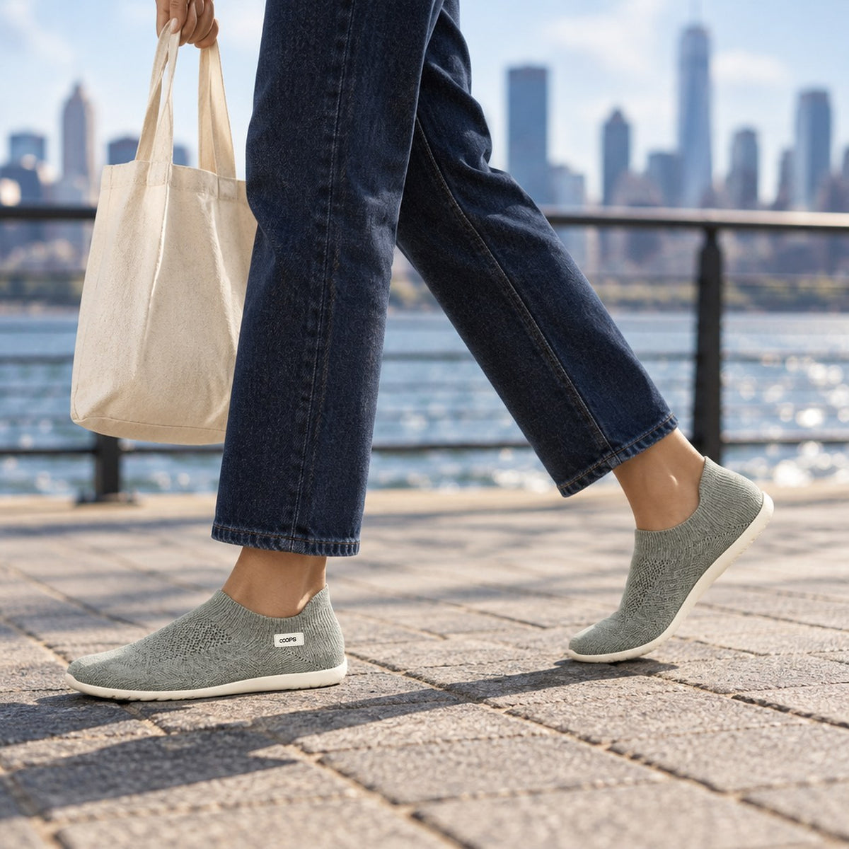 A person walking a city waterfront path in Ash Gray COOPS sock shoes with a tote bag