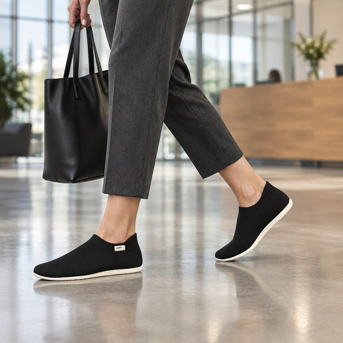 A person walking through an office lobby in Black COOPS sock shoes and tailored trousers