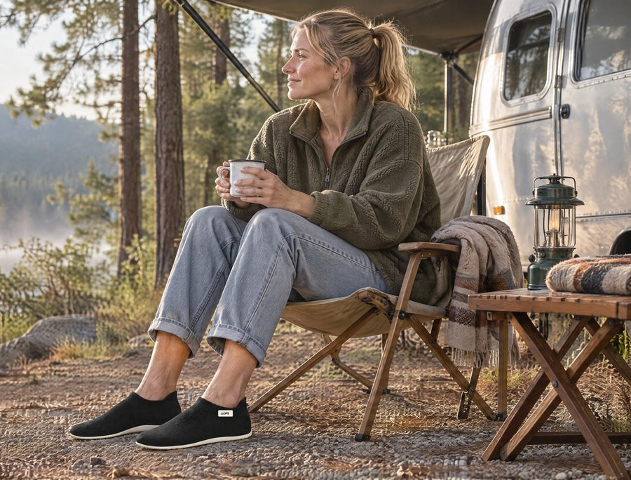 Man with a coffee beside a travel trailer in a pine forest, wearing Sand COOPS Low Top sock shoes