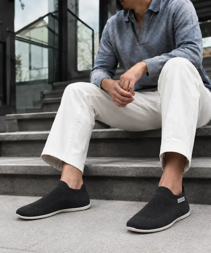 A man sitting on concrete steps in white trousers and Black COOPS Low Top sock shoes