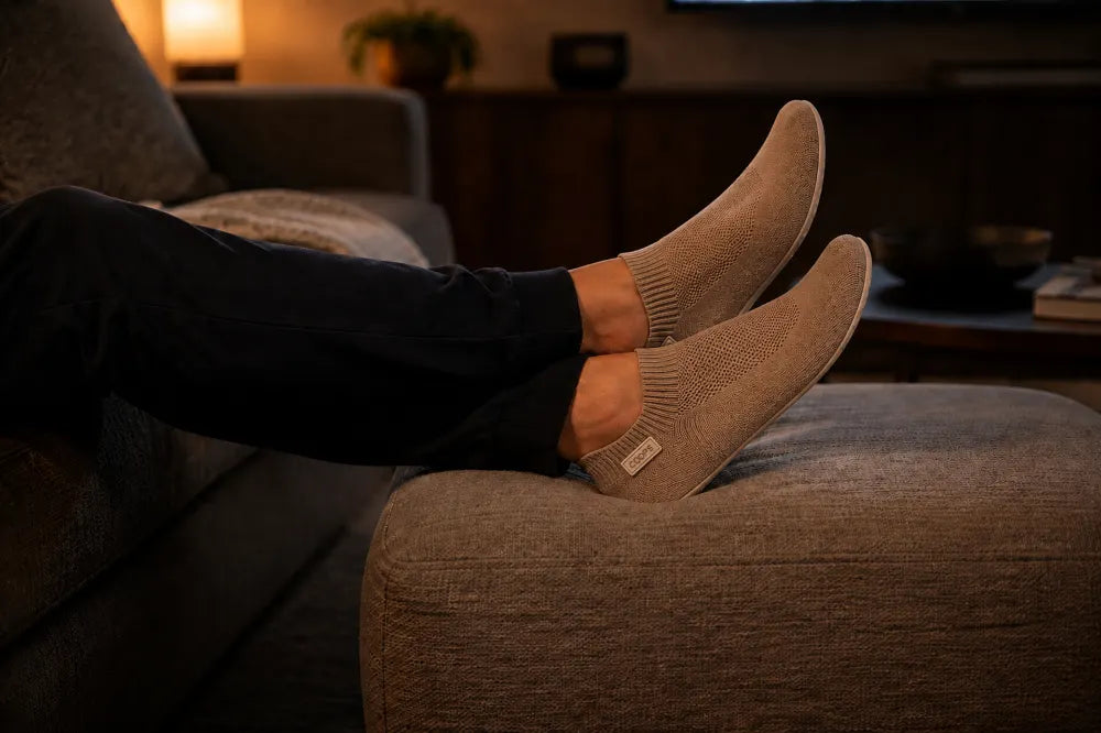 Feet in sand COOPS Low Tops resting on an ottoman in front of a television.