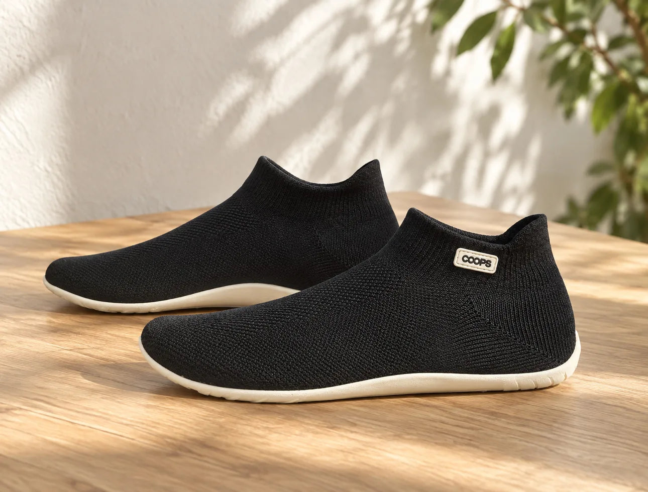 A pair of Black COOPS sock shoes on a wooden table in soft sunlight, a plant behind